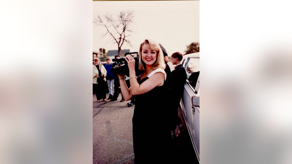 Jodi Huisentruit holding a camera wearing a black dress and smiling.