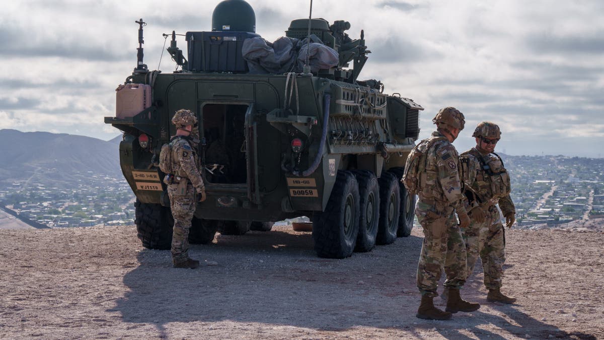 U.S. soldiers in combat gear stand beside a military armored vehicle on a hill overlooking a city, with mountains and overcast skies in the background.