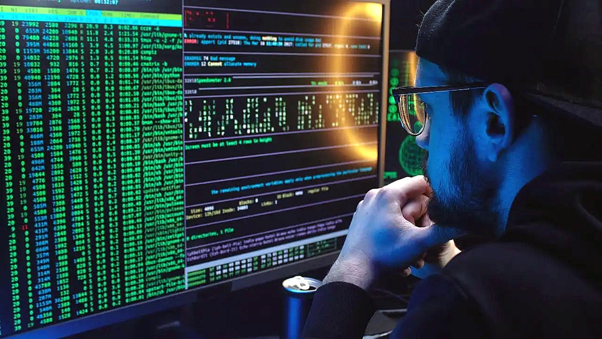 A man stares at computer code on hiis monitor in a darkened room, with a ring light reflected on the screen and an open canned beverage next to him.