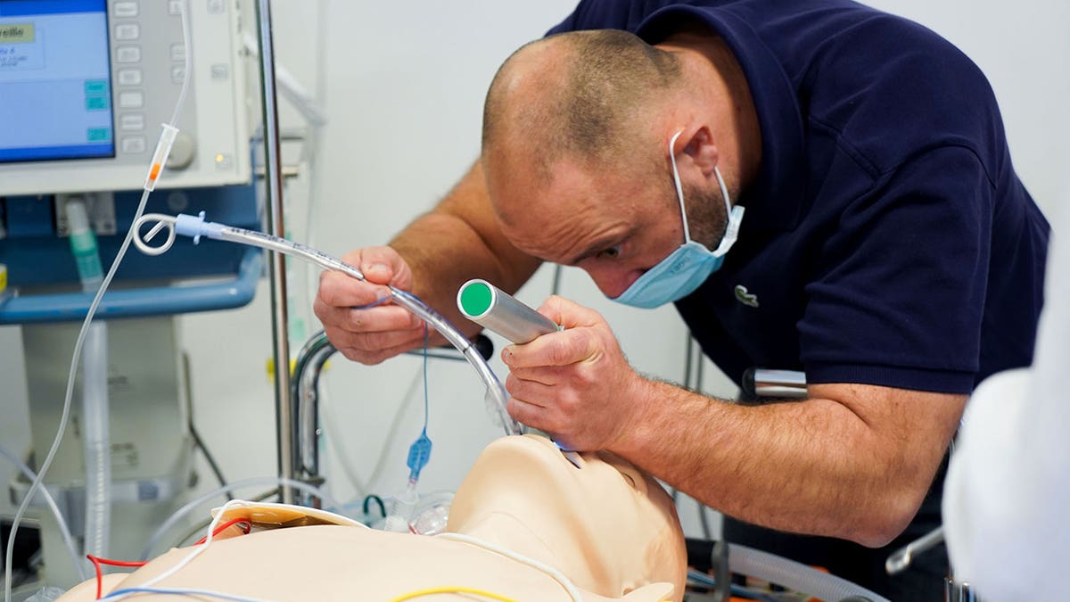 Medical worker practices intubation on a training mannequin in a hospital simulation room.