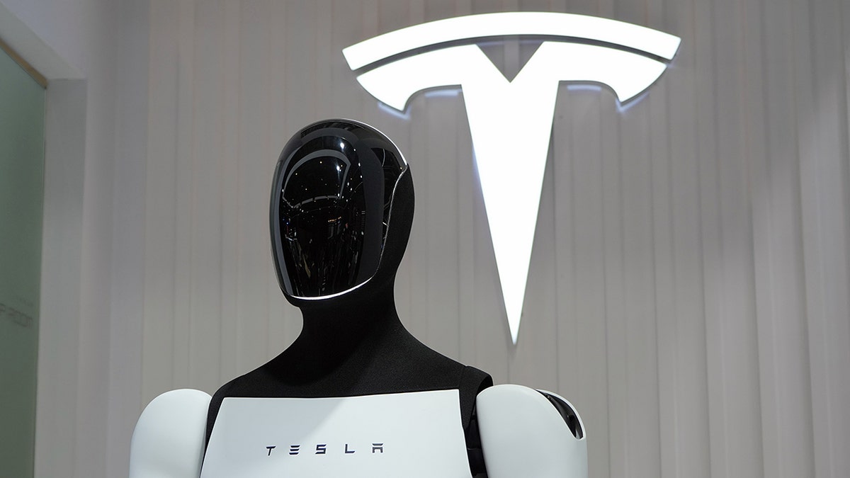 A Tesla Optimus robot with the Tesla logo behind it