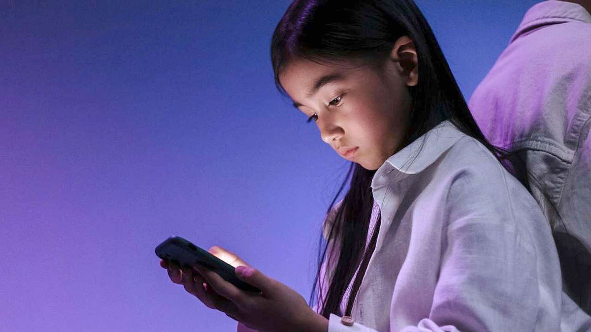 A girl looks at a smartphone in front of an indigo background.
