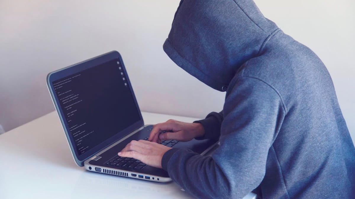 Someone with a hoodie types suspiciously on a laptop that displays a dark screen.